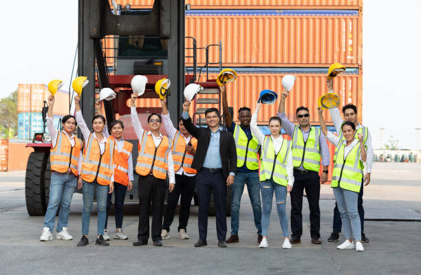 portrait factory workers or engineers team holding helmet and raise hand up for success in containers warehouse storage